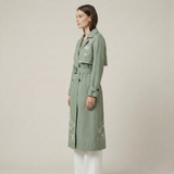 Woman wearing a green trench coat with floral embroidery on a plain background