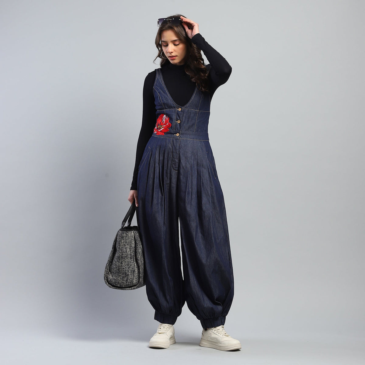 Woman wearing a denim jumpsuit with a red flower design, holding a gray bag against a plain background.