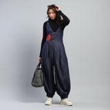 Woman wearing a denim jumpsuit with a red flower design, holding a gray bag against a plain background.